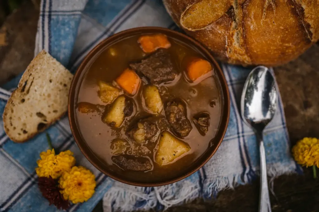 A dark bowl of beef stew with carrots beside slices of sourdough bread