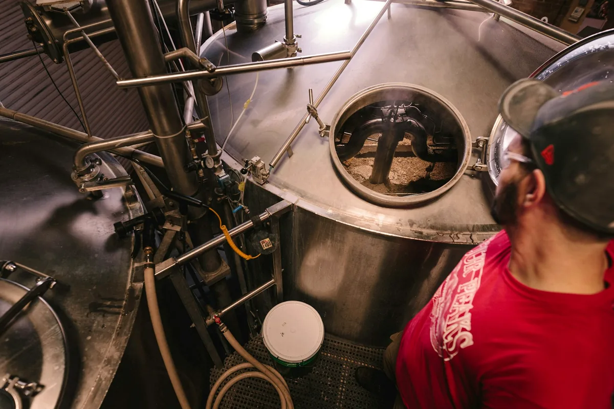 A brewer leaning over the open hatch of a large brewing vessel