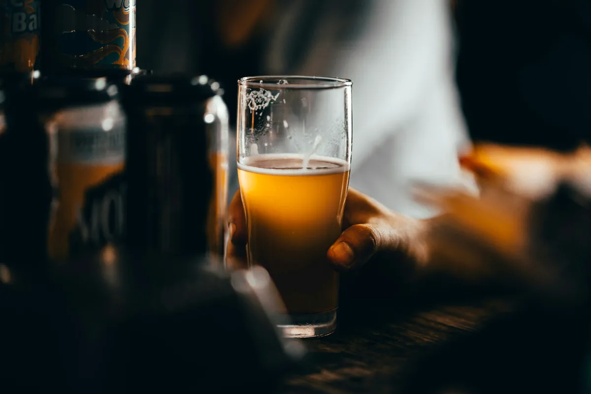 A hand holding a glass of pale beer at a dimly lit bar