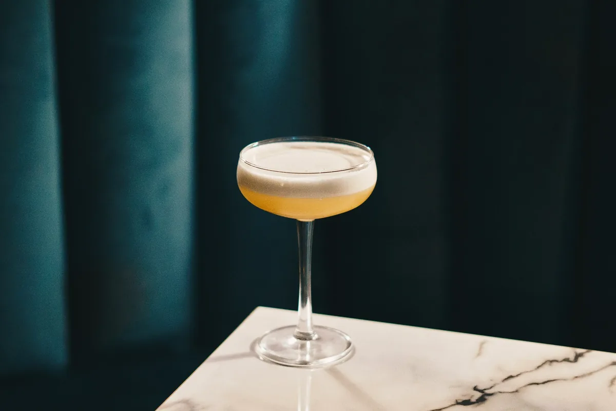 A pale sour cocktail with a foam top in a stemmed coupe on marble