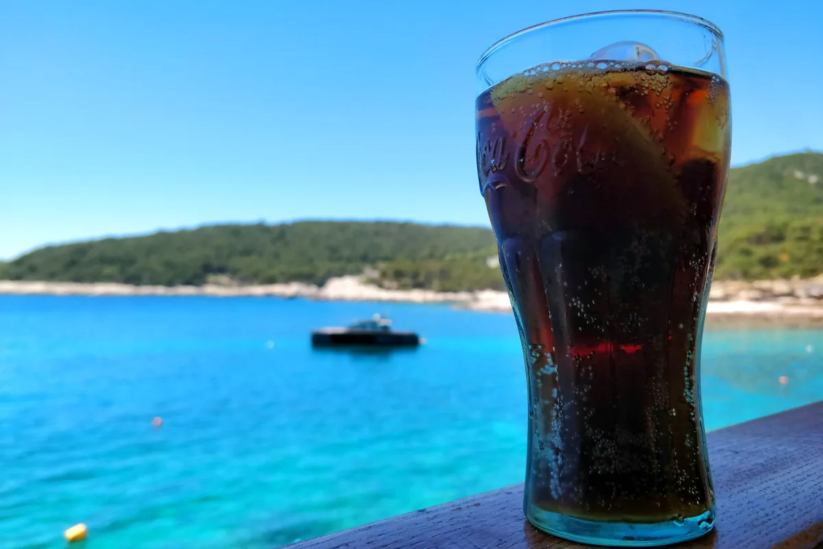 A tall glass of cola over ice on a table with the sea behind