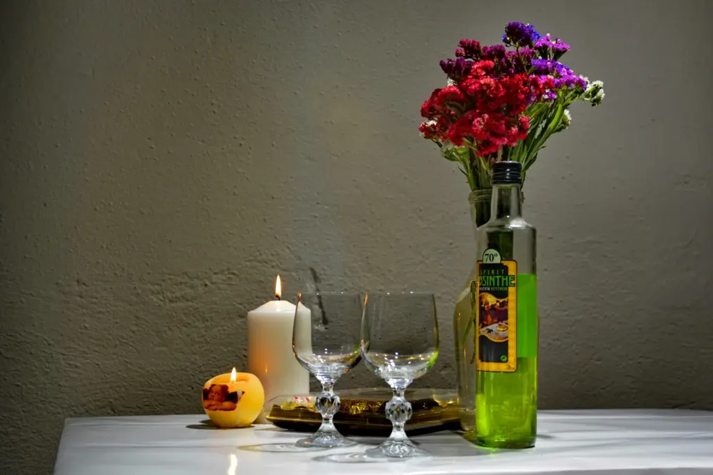 A green absinthe bottle beside two small stemmed absinthe glasses and a candle