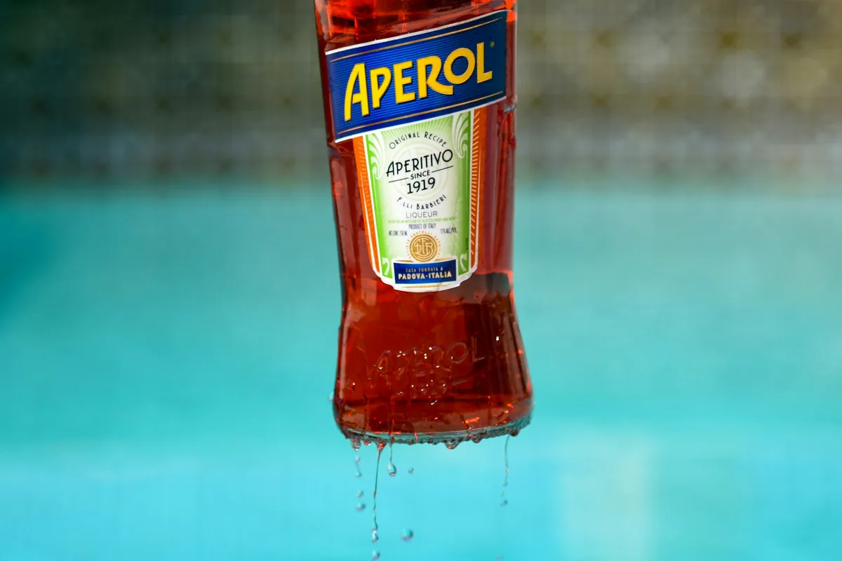 A bottle of Aperol lifted dripping above bright blue water
