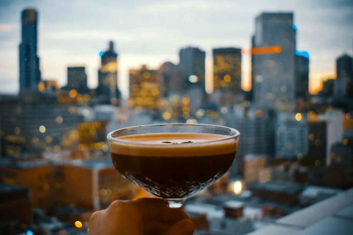 An espresso martini held up against a city skyline at dusk