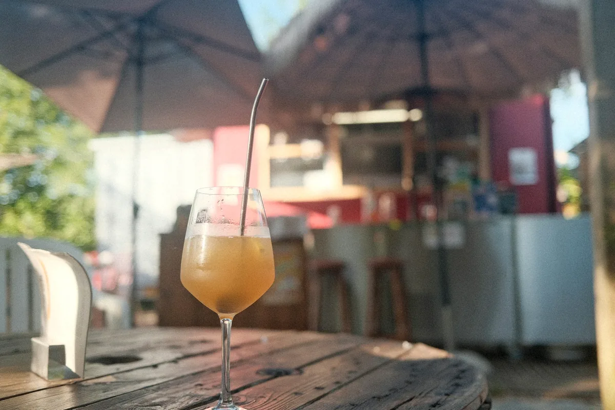 A tall orange drink on a wooden table under a parasol at an outdoor cafe