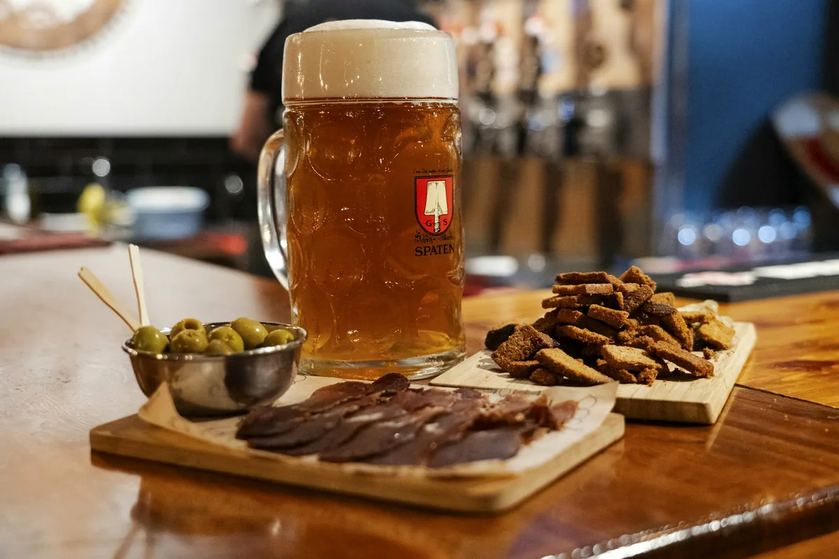 A wooden board of snacks and charcuterie beside a tankard of beer in a bar