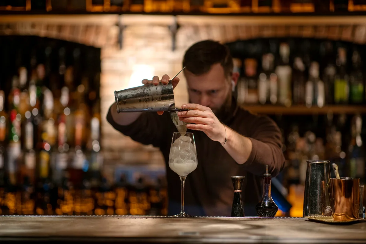 A bartender straining a drink at a lit back bar lined with bottles