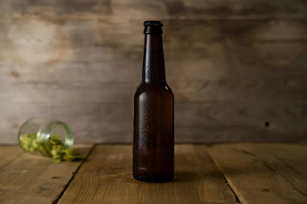 A brown beer bottle on a wooden table with hop cones spilt behind it
