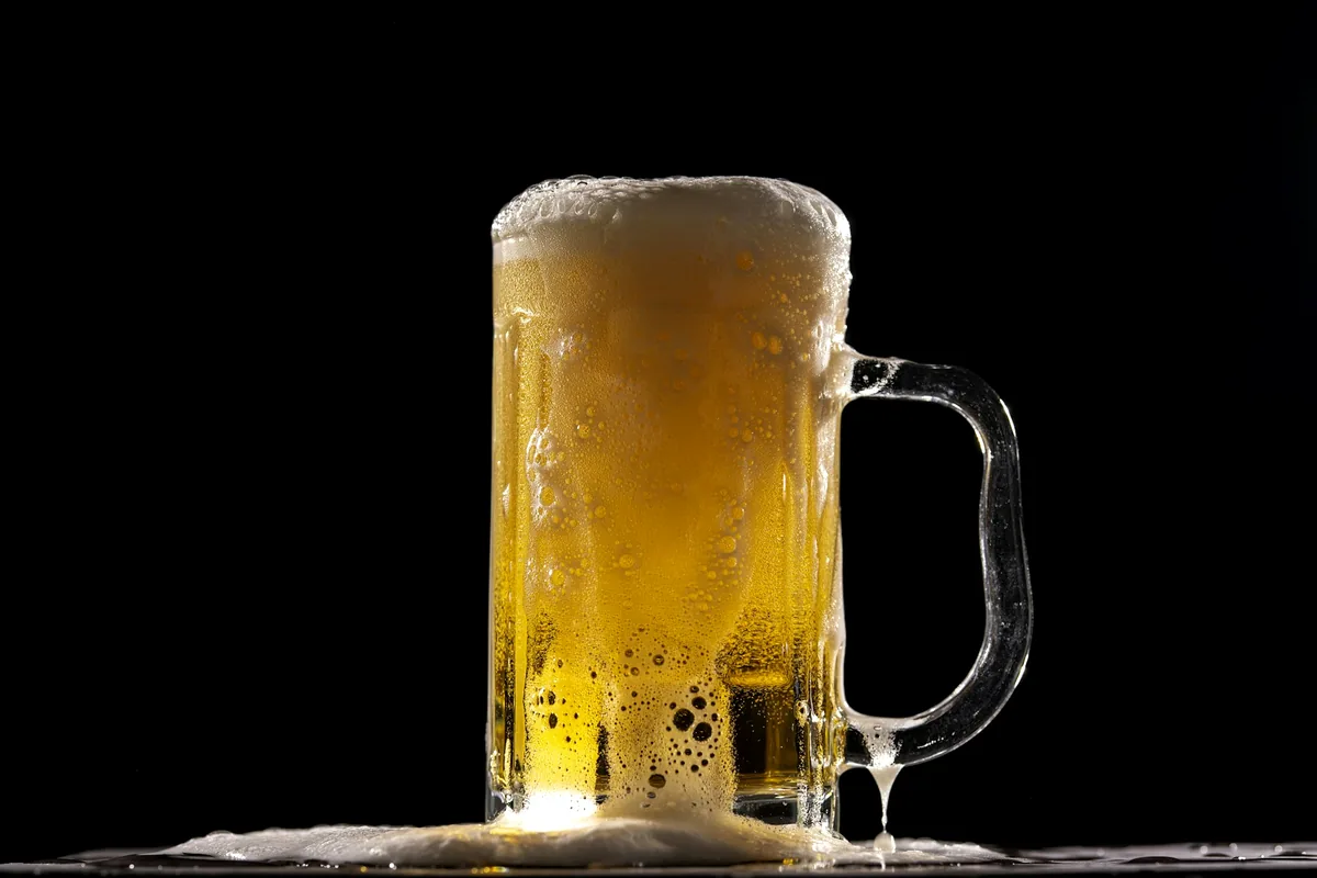 A glass tankard of pale beer with a thick foam head on a black background