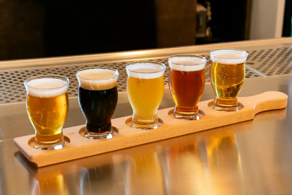 Five small glasses of beer in a wooden flight paddle, ranging from pale gold to near black