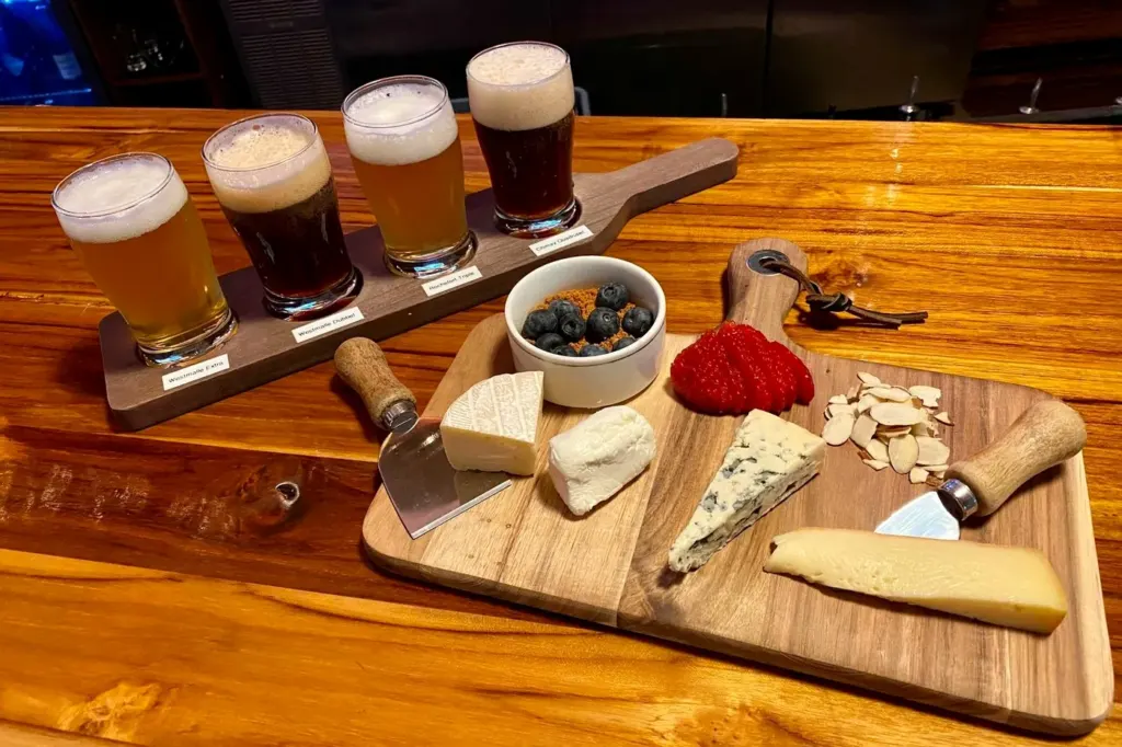 A flight of four Belgian beers beside a wooden board of cheeses, nuts and preserves