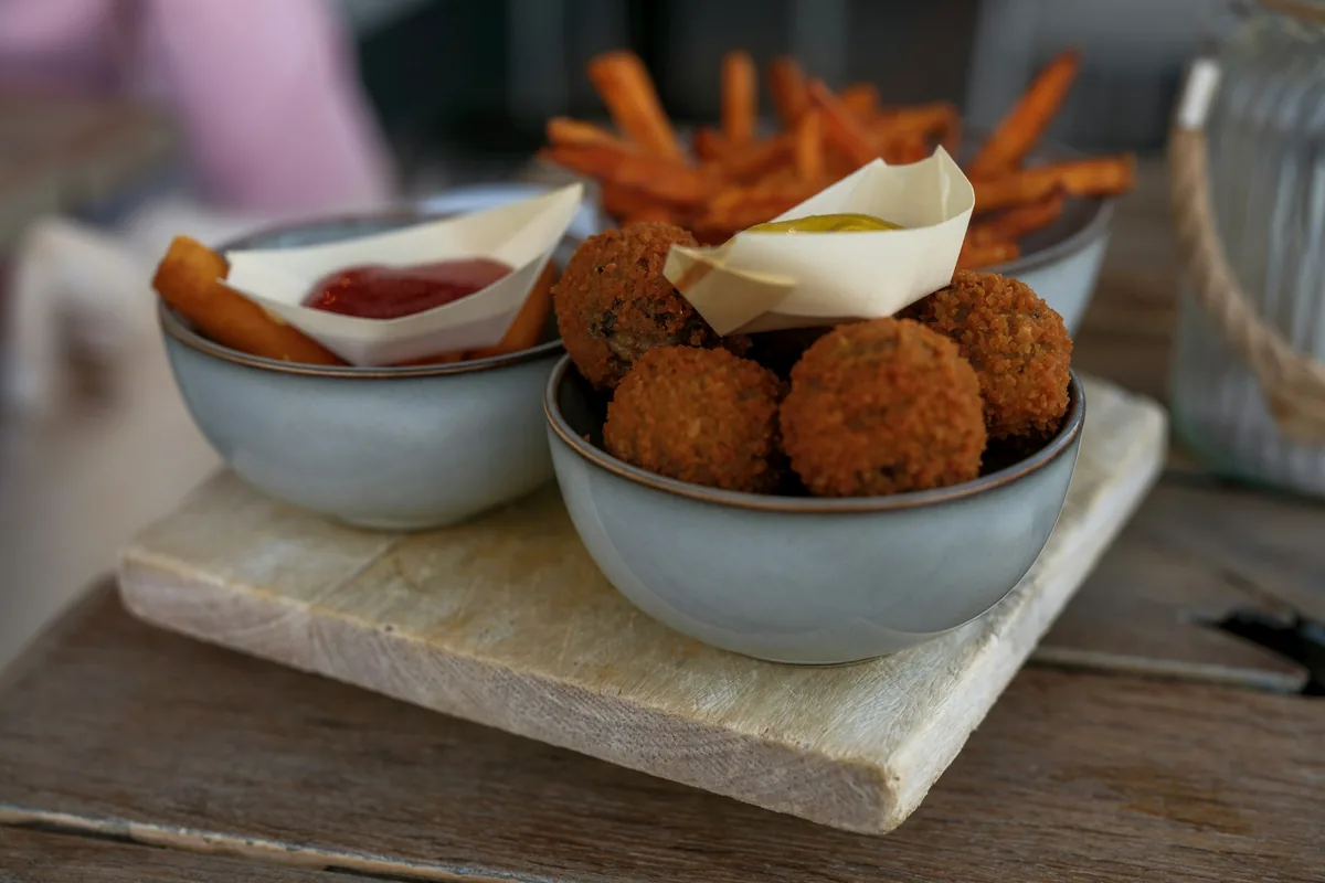 Two bowls of bitterballen beside mayonnaise and sweet potato fries on a wooden board