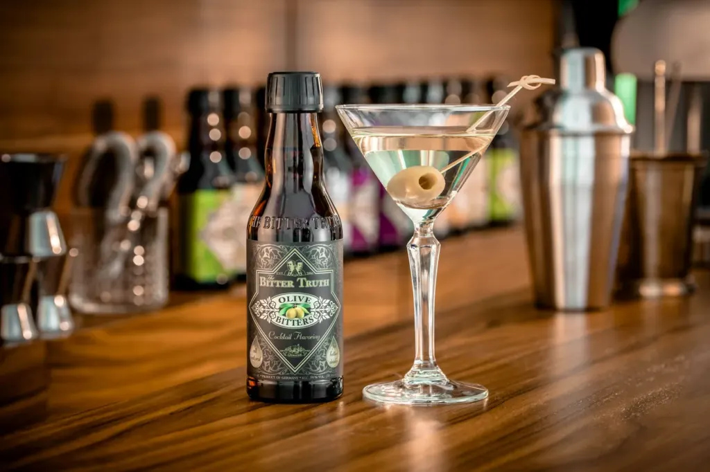 A small dark bitters bottle on a wooden bar beside a martini glass and a shaker