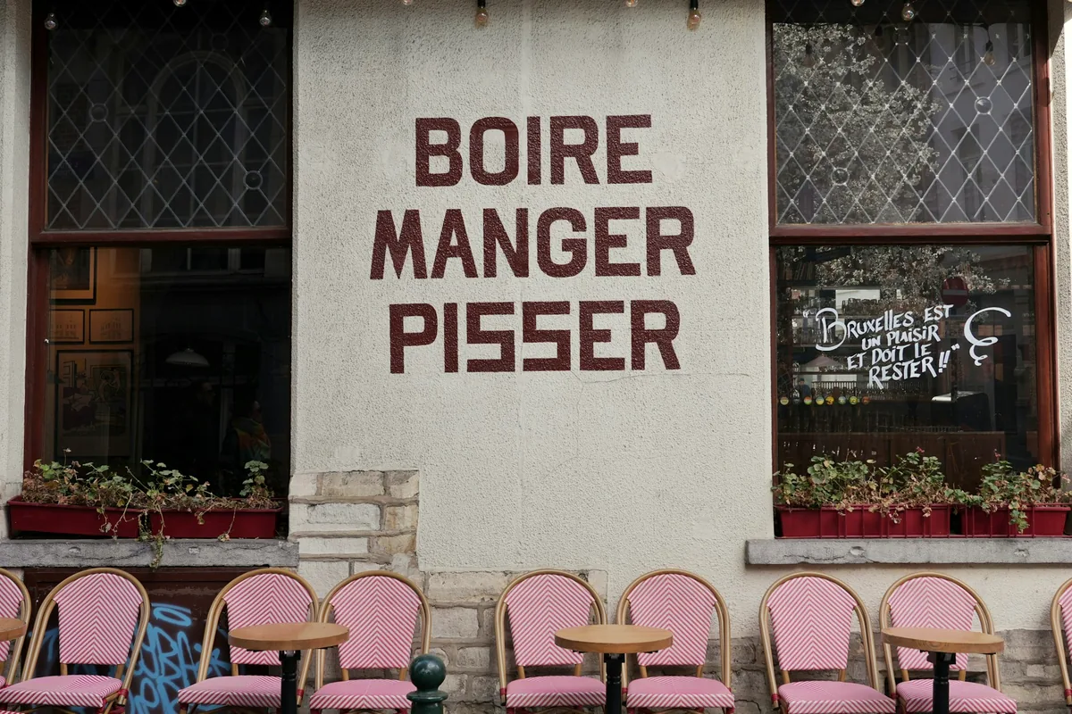 A cafe frontage in Brussels painted with the words boire, manger, pisser above pink chairs