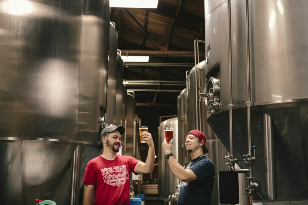 Two brewers tasting beer from glasses beside stainless fermentation tanks