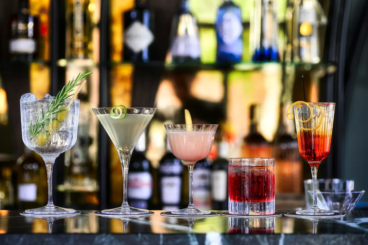 A row of finished cocktails in assorted glassware along a lit back bar