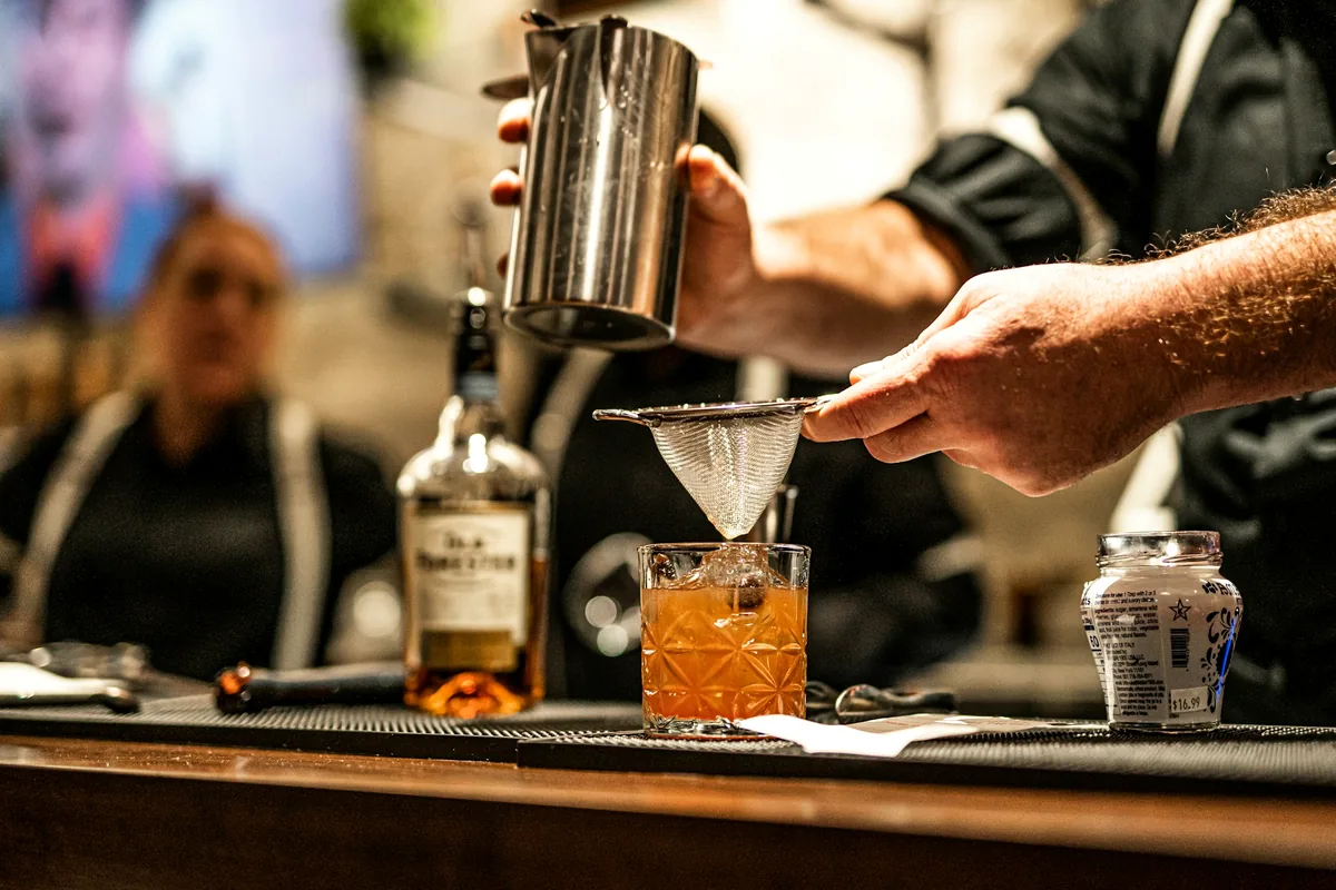Hands straining a cocktail through a fine strainer into a stemmed glass on a dark bar