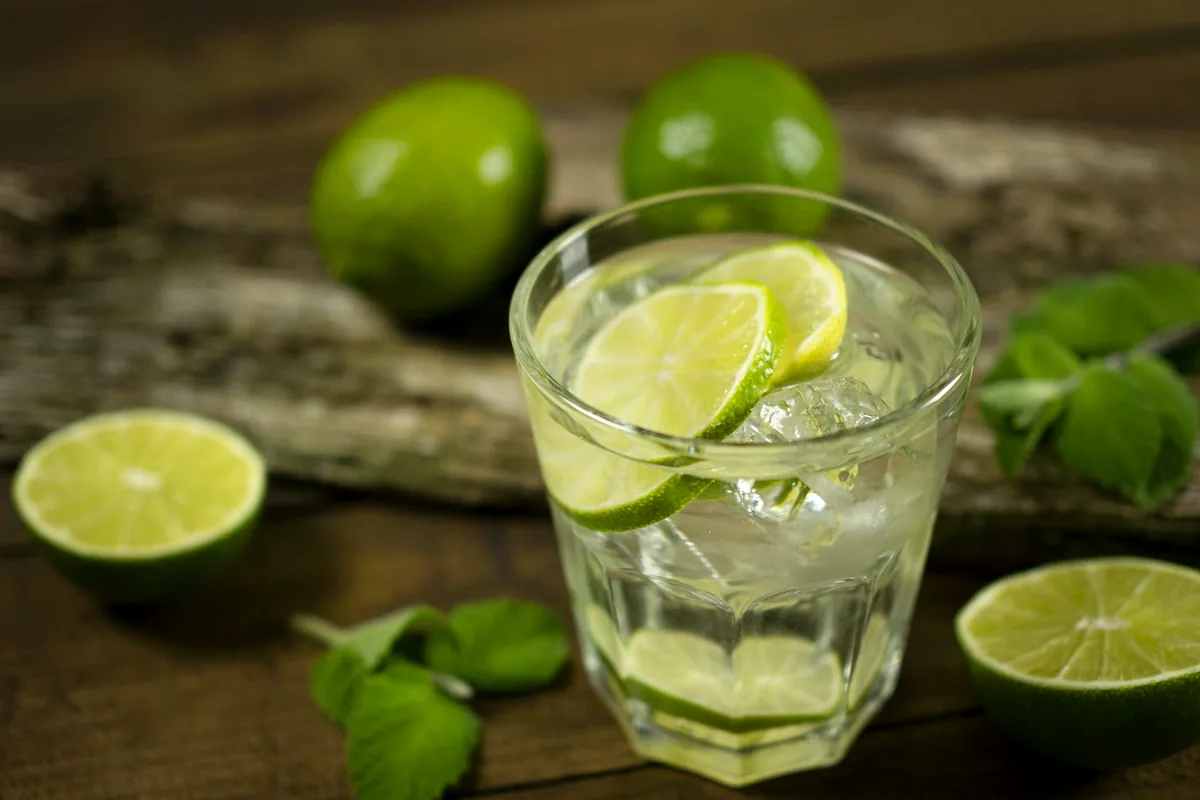 A glass of clear drink with lime wedges and whole limes on weathered wood