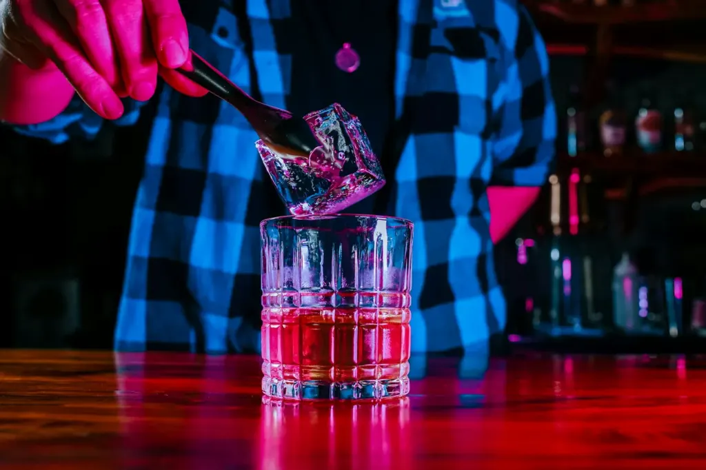 Tongs lowering a large clear ice cube into a tumbler at a bar