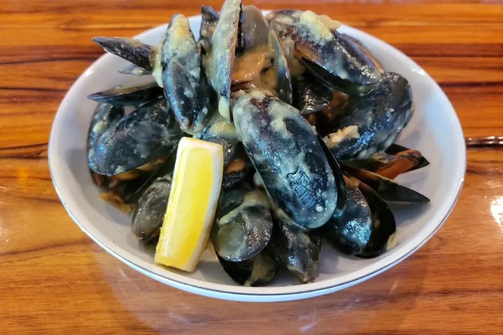 A white bowl of steamed mussels with a lemon wedge on a wooden table