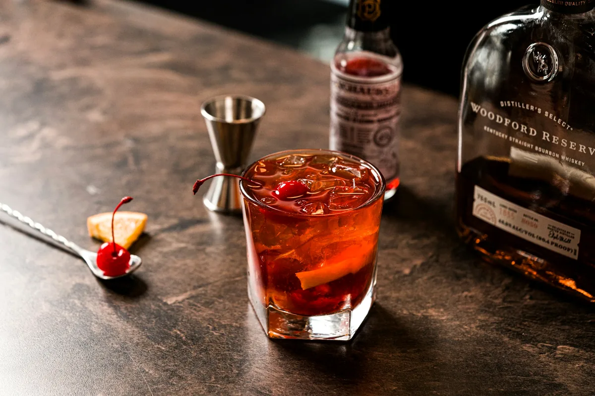 An amber cocktail over ice with cherries and orange peel, a jigger and a whiskey bottle behind