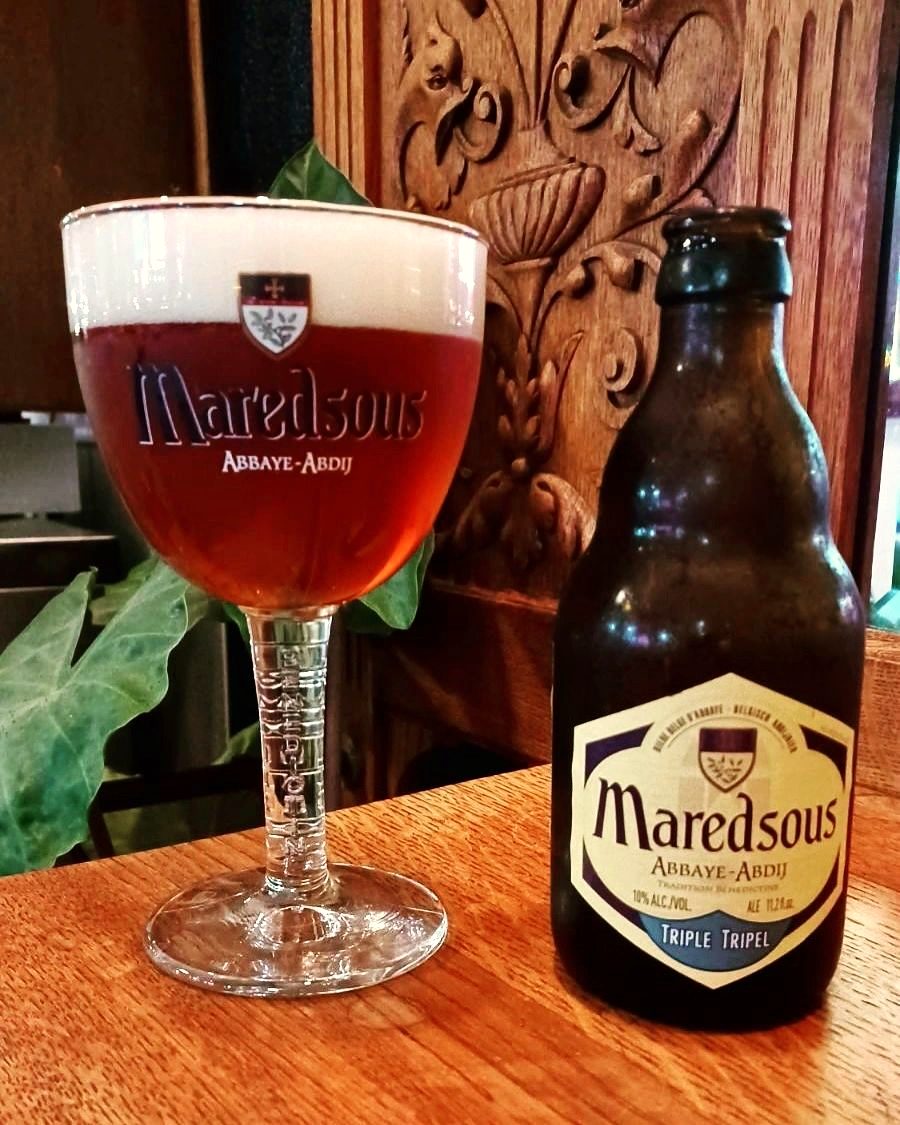 A Maredsous abbey chalice of amber Belgian ale beside its bottle, against carved wood