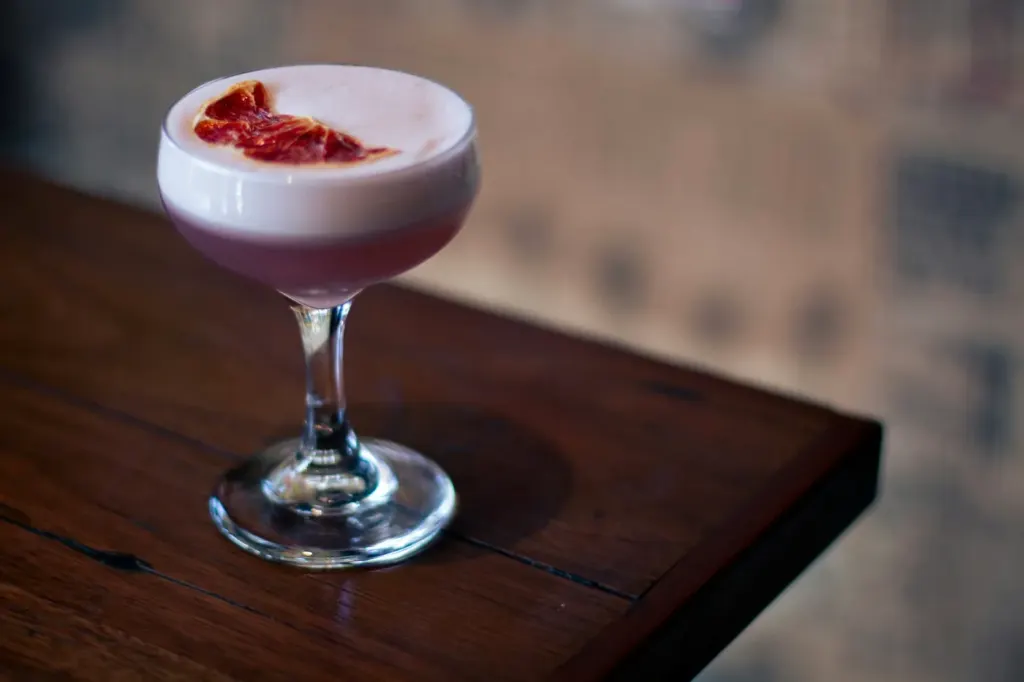 A pale pink cocktail with a thick foam top in a coupe on a dark table