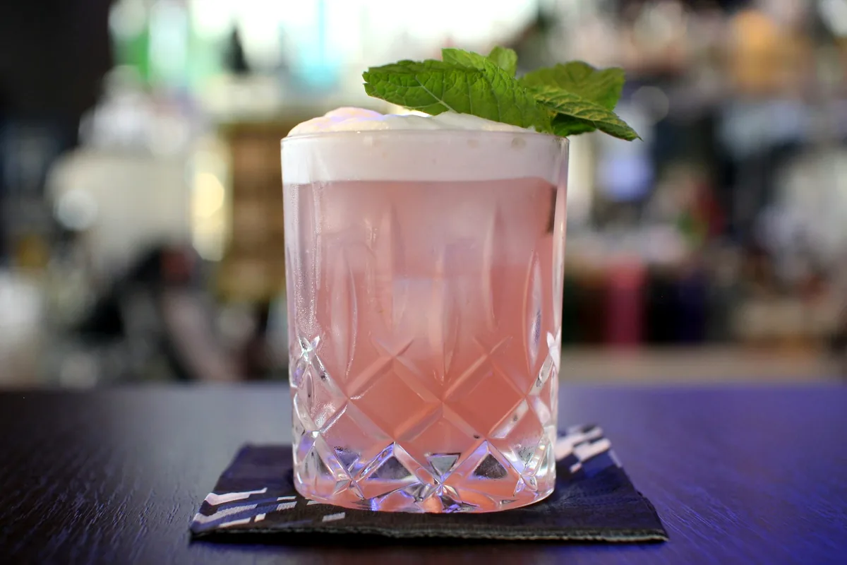 A pink foam-topped drink in a cut glass with a sprig of mint