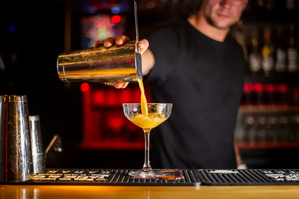 A bartender pouring a golden drink from a shaker into a coupe glass