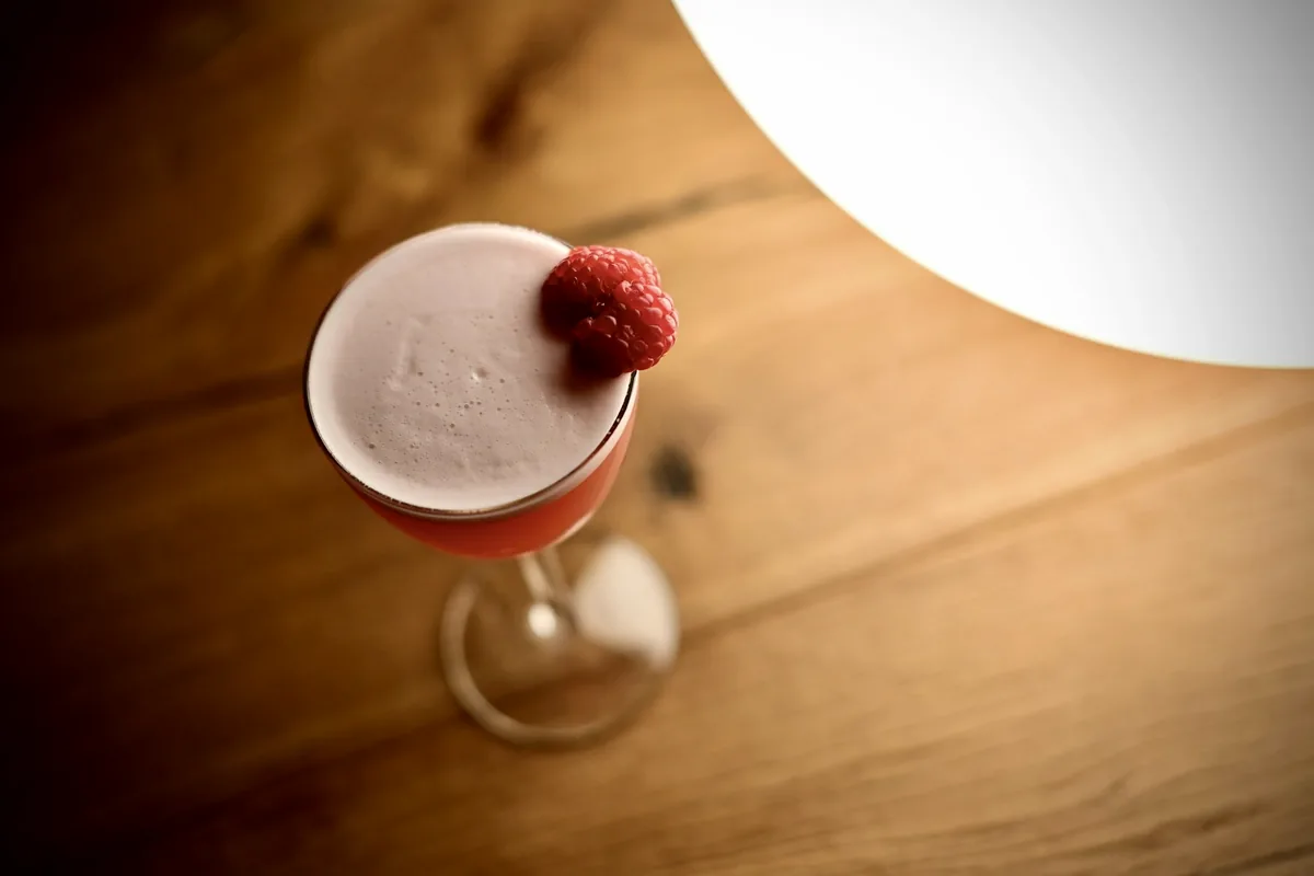 A pink cocktail with a single raspberry balanced on the rim