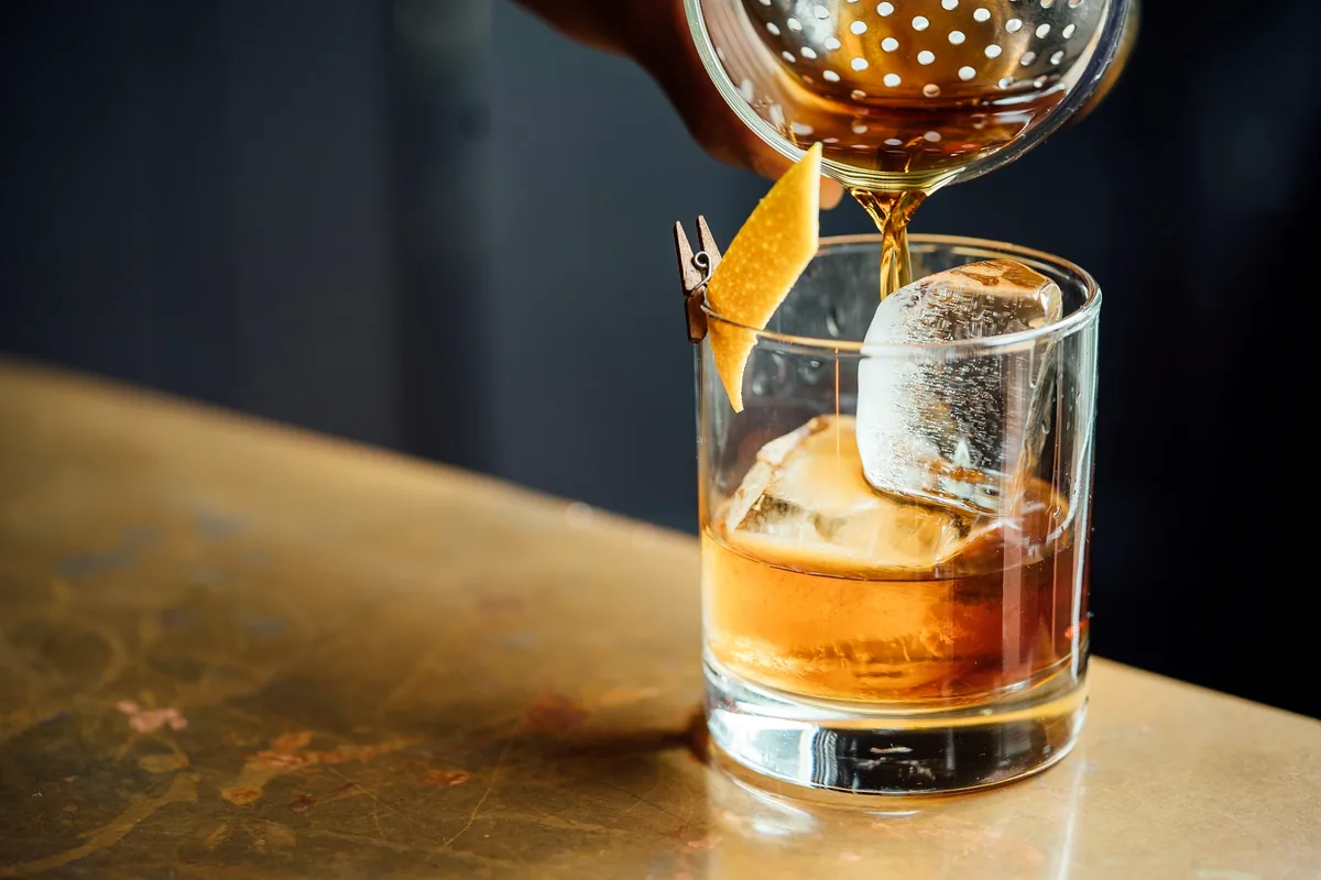 A dark amber cocktail being strained over a large ice cube with a citrus twist