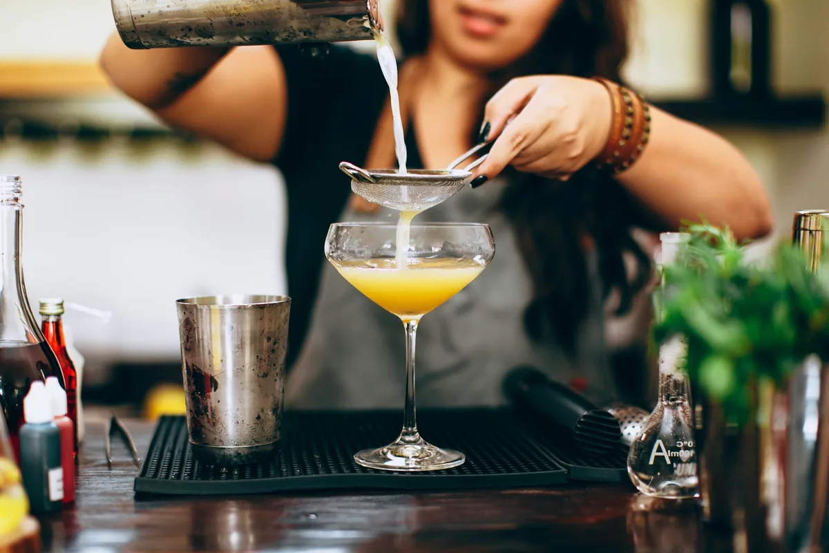 A bartender straining a pale cocktail through a strainer into a stemmed glass