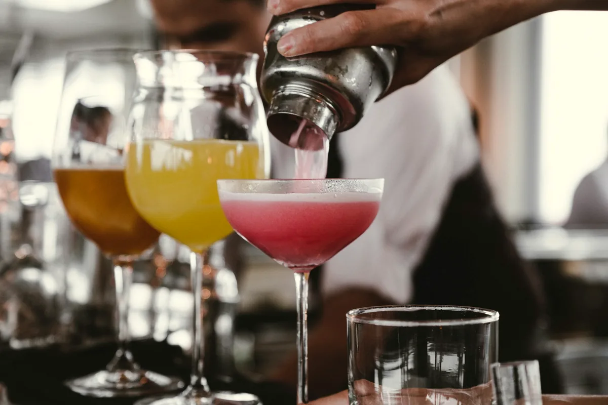 A bartender straining a pink cocktail into a chilled coupe beside a row of filled glasses
