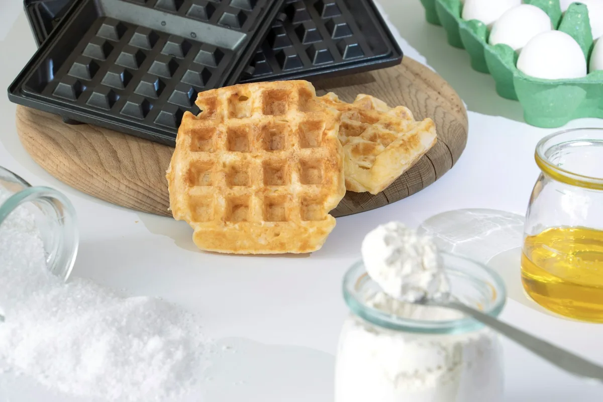 A waffle maker with cooked waffles, eggs and flour on a counter