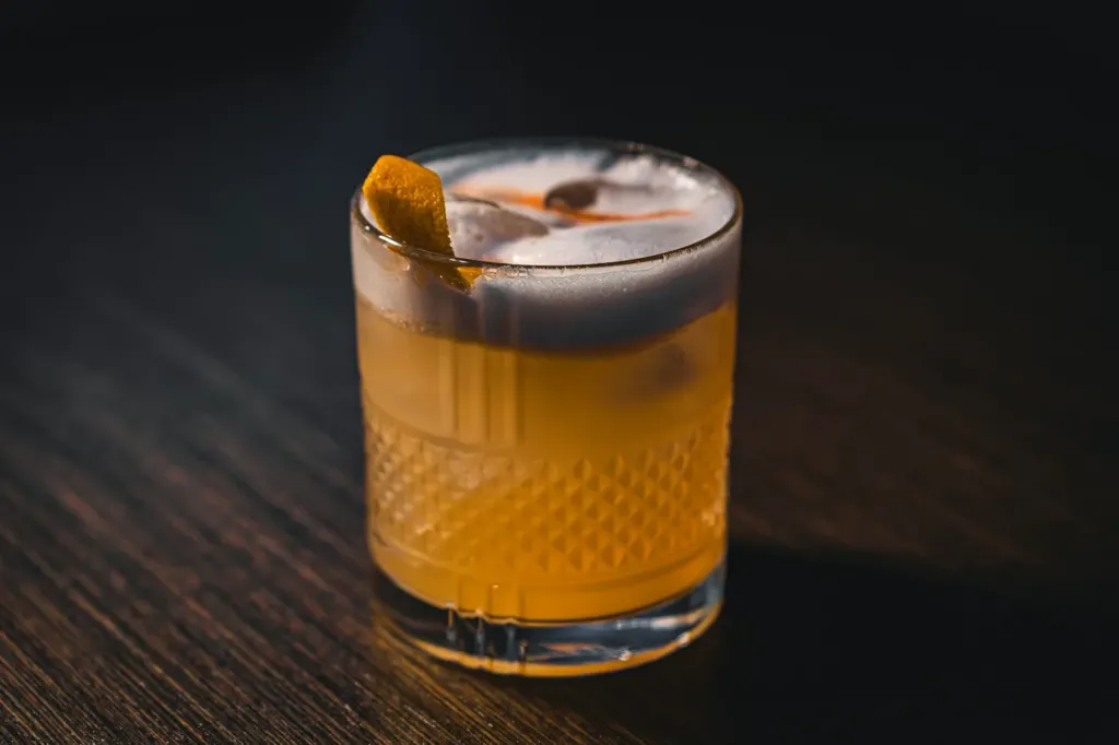A whiskey sour in a rocks glass with a thick pale foam cap and an orange twist